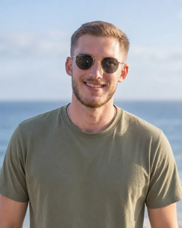 Portrait d’Anthony Decker, souriant, avec des lunettes de soleil et un t-shirt kaki, devant la mer.